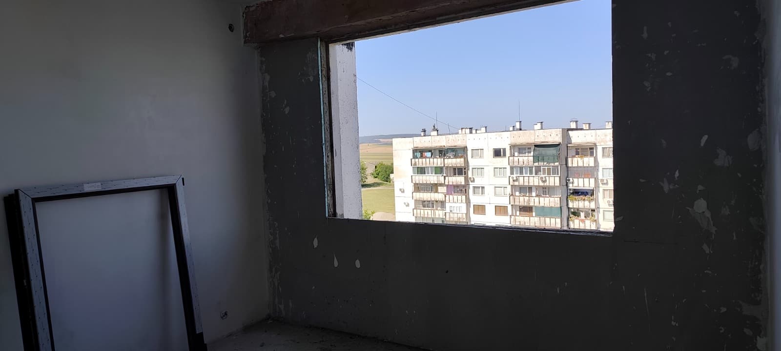 Празен отвор за прозорец в стена с тъмна мазилка; до съседната стена е подпряна рамка
