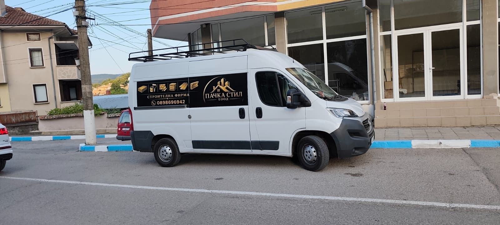 Белият бус на Пачка Стил с логото на фирмата и телефона, спрял пред сграда