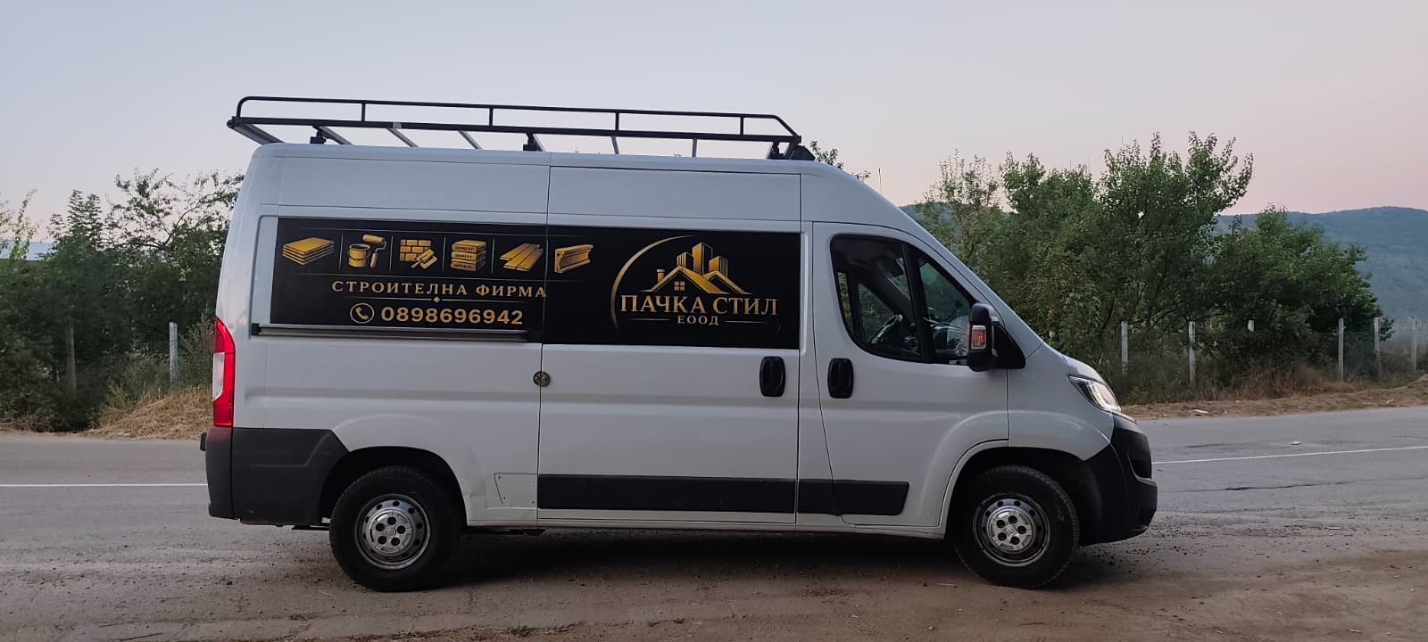 Бял бус на Пачка Стил, сниман отстрани по здрач: на борда са логото, надпис „Строителна фирма“ и телефон 0898 696 942
